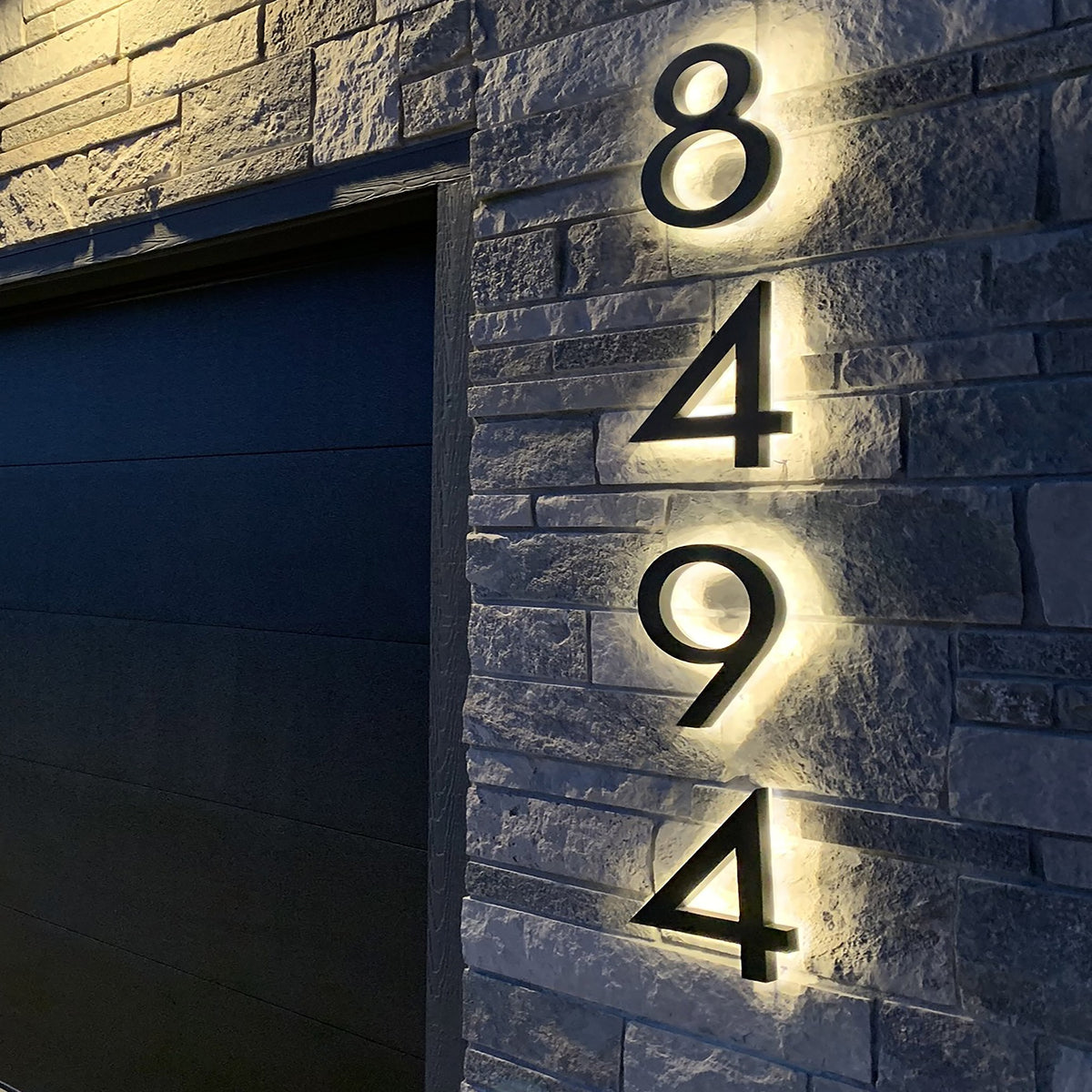 Black backlit LED house numbers 8494 lit on a stone wall